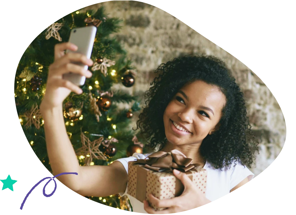 A woman taking a selfie by a Christmas tree holding a gift